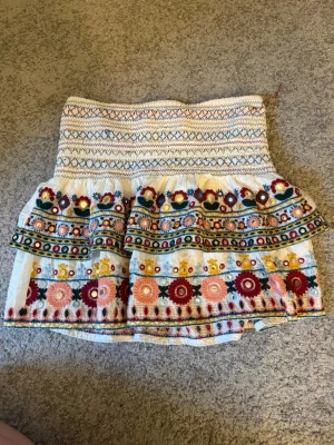 Vit kortkjol med broderade blommor -  vit kortkjol med färgglada broderade blommor. Har volang och kan användas som en topp också.