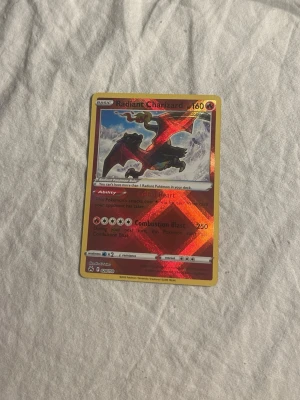 Radiant Charizard Pokémonkort - Säljer ett Radiant Charizard Pokémonkort med holografisk effekt. Kortet har en färgstark design med röd, gul och svart färg och visar Charizard i en actionfylld pose. Perfekt för samlare eller Pokémonfans.