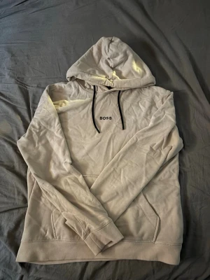 Beige hoodie från BOSS med huva - Snygg beige hoodie från BOSS med svart dragsko i huvan och broderad BOSS-logga på bröstet. Hoodien har känguruficka framtill och är tillverkad i mjukt material som känns skönt mot huden. Perfekt för en avslappnad och trendig look.