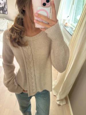 Beige stickad tröja med flätmönster - Mysig beige stickad tröja med rund halsringning och ett snyggt flätmönster framtill. Tröjan har långa ärmar och ribbade muddar vid ärmslut och nederkant. Perfekt för lager på lager och passar till jeans eller kjol.