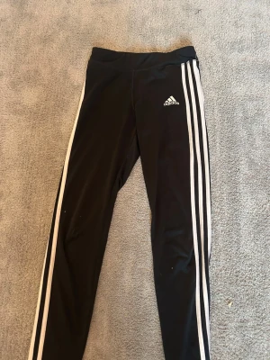 Svarta Adidas leggings med vita ränder - Säljer ett par svarta Adidas leggings med klassiska vita ränder längs benen och Adidas-logga på låret. Tillverkade i Climalite-material som andas och håller dig torr. Perfekta för träning eller chill dagar. Tight passform och elastisk midja. Jag har storlek XS men kan ha dessa🤍Pris diskuteras gärna då jag vill bli av med grejer