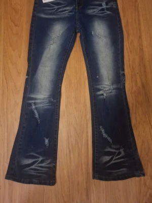 Blå bootcut jeans med slitningar - Säljer ett par blå bootcut jeans med tydliga slitningar och blekta detaljer på benen. Jeansen har en klassisk femficksmodell och är tillverkade i denim med stretch. Perfekta för en avslappnad och trendig look.