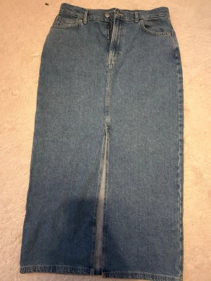 Lång jeanskjol med slits framtill - Säljer en klassisk blå lång jeanskjol i rak modell med slits framtill. Kjolen har femficksdesign, bälteshällor och stängs med knapp och dragkedja. Perfekt till sneakers eller boots för en cool vardagslook.