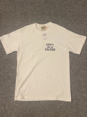 Vit t-shirt från Gallery Dept - Vit t-shirt från Gallery Dept med trycket 'DÉPT. de la GALERIE' i svart på både bröstet och ryggen. Klassisk passform och rund halsringning. Tillverkad i mjuk bomull, perfekt för en clean och stilren look.