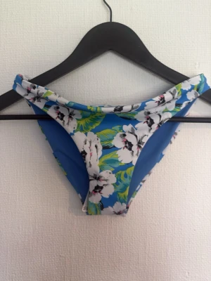 Blå blommig bikiniunderdel - Säljer en snygg bikiniunderdel i blått med stort blommigt mönster i vitt, svart och grönt. Klassisk modell med medelhög midja och stretchigt material som sitter skönt. Perfekt för strandhäng eller pooldagar!