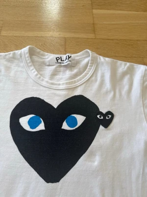 CDG T shirt  - Säljer en vit t-shirt från Comme des Garçons PLAY med det ikoniska svarta hjärtat med blå ögon tryckt på bröstet. Klassisk rund hals och korta ärmar. Perfekt t shirt för dig som gillar streetwear och cool design. Plagget är i storlek S och något liten i storlek, mycket bra skick. 
