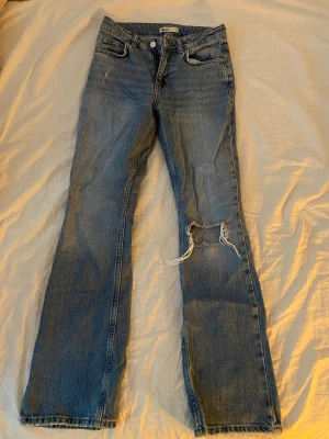Blå slitna jeans från g perfect jeans - Ett par blå jeans från g perfect jeans med raka ben och slitningar, inklusive ett stort hål på ena knät. Klassisk femficksmodell och knappgylf. Jeansen har en avslappnad passform och är tillverkade i denim.