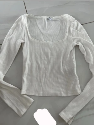 Vit ribbad långärmad topp - Snygg vit ribbad topp med lång ärm och fyrkantig urringning. Dekorativ spetskant runt halsen ger en söt touch. Perfekt att styla med jeans eller kjol för en trendig look.