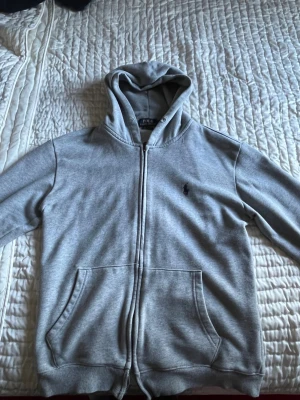 Grå hoodie från Polo Ralph Lauren - Säljer en grå hoodie från Polo Ralph Lauren med dragkedja, huva och fickor framtill. Klassisk logga broderad på bröstet. Tillverkad i mjukt bomullsmaterial som är skönt att ha på sig. Perfekt för en avslappnad och stilren look.