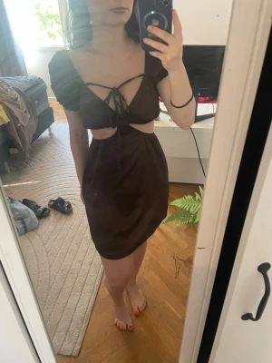 Brun klänning med cut-outs och knyt - Snygg brun klänning med kort ärm, cut-outs vid midjan och knyt framtill. Klänningen har en figurnära passform och är perfekt för dig som vill sticka ut. Materialet känns lätt och bekvämt mot huden.