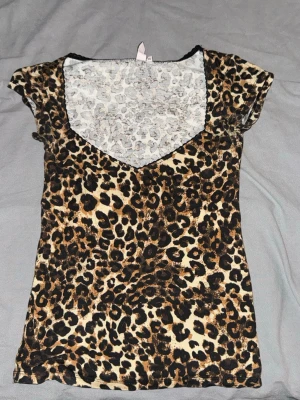 Leopardmönstrad topp från Nelly - Snygg leopardmönstrad topp från Nelly med v-ringning och korta ärmar. Tunn och stretchig, perfekt för dig som vill sticka ut. Djurmönstret går i beige, brunt och svart. Passar till både jeans och kjol för en trendig look. Aldrig använd!