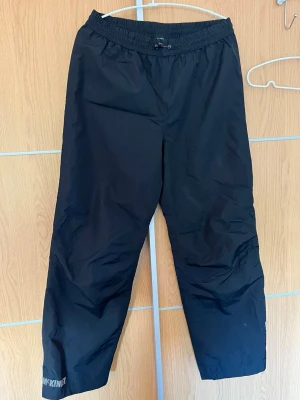 Svarta täckbyxor från McKinley - Svarta täckbyxor från McKinley med elastisk midja och dragsko. Perfekta för kalla dagar och utomhusaktiviteter. Tillverkade i slitstarkt material som står emot väta och vind. Snygg och enkel design med logga på benet.