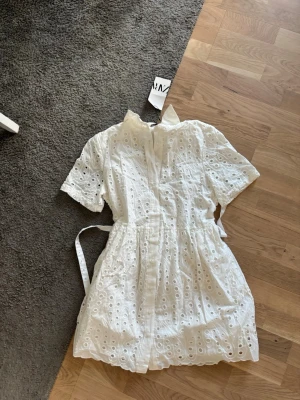 Vit kort klänning - Söt vit kortklänning med broderade hål och knytband i midjan. Perfekt nu till sommaren! Aldrig använd :)