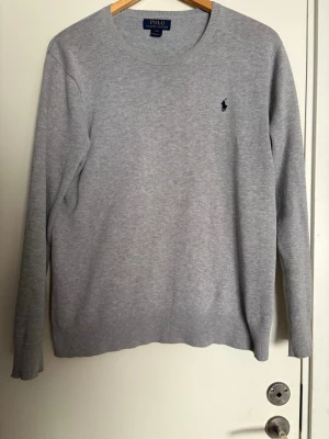 Ljusgrå stickad tröja Polo Ralph Lauren - Snygg ljusgrå stickad tröja från Polo Ralph Lauren med rund halsringning och diskret broderad logga på bröstet. Tröjan har långa ärmar och ribbade muddar vid ärmslut och nederkant. Perfekt till lager på lager och enkel att matcha.