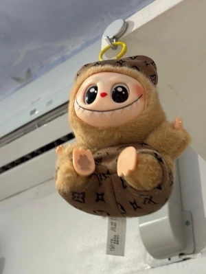 Labubu - En gullig mjuk plyschfigur med stort leende och stora ögon. Figuren har beige päls, beige ansikte och tassar samt brun overall med svart mönster. Perfekt som dekoration eller nyckelring. Kan hängas upp med en gul krok.