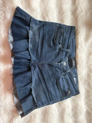 Mörkblå jeanskjol med volang - Säljer en mörkblå jeanskjol med råa kanter och volang nedtill. Kjolen har klassiska fickor fram och bak samt knapp och dragkedja i midjan. Perfekt för dig som gillar en edgy och trendig stil.