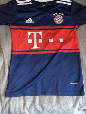 Bayern München matchtröja Adidas - Snygg Bayern München matchtröja från Adidas i mörkblått med röda och vita detaljer. Tröjan har klubbmärke, Adidas-logga och Telekom-sponsor på bröstet samt vita ränder på axlarna. Tillverkad i lätt och ventilerande climacool-material.