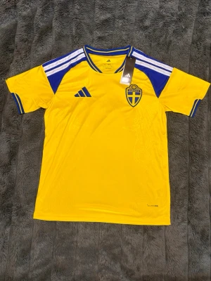 Sverige VM fotbollströja Adidas gul/blå - Gul VM fotbollströja från Adidas med blå detaljer och Sveriges landslagsemblem på bröstet. Tröjan har korta ärmar, rund hals och vita ränder på axlarna. Vid frågor är det bara skriva och priset är varken hugget i sten, finns 2st