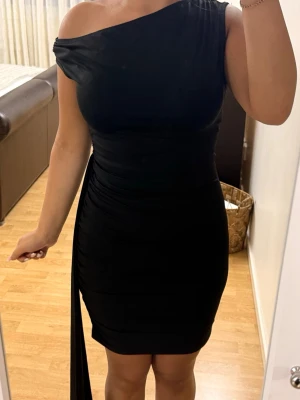 Svart one shoulder kort klänning - Snygg svart kort klänning med one shoulder-design och draperad detalj på sidan. Klänningen har en figurnära passform och är tillverkad i ett stretchigt material som sitter bekvämt. Perfekt för dig som vill sticka ut med en stilren look. Jätte fint skick, minns dock ej vart den är köpt ifrån, priset kan diskuteras💕