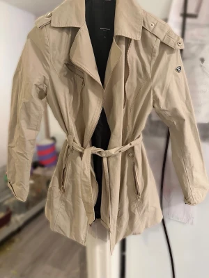 Trenchcoat med bälte RockAndBlue S/36 - Stilren beige trenchcoat med klassisk krage, axelklaffar och bälte i midjan. Jackan har dragkedja framtill, två fickor med dragkedja och ett emblem på ärmen. Storlek S/36 från Rockandblue. Liten defekt förekommer, se sista bild. Annars fint skick!