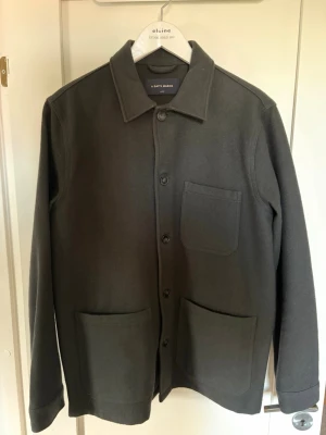 Mörkgrön ull overshirt från A Day's March - Stilren mörkgrön ull overshirt från A Day's March med klassisk krage och tre utanpåliggande fickor. Overshirten har raka ärmar och knäppning framtill. Perfekt lager-på-lager-plagg i ett mjukt och tjockare material som ger en clean look.