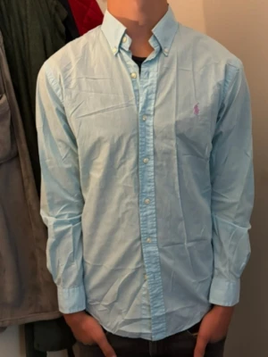Ljusblå skjorta med knappar - Snygg ljusblå skjorta med klassisk krage och knappar hela vägen framtill. Skjortan har lång ärm och en liten broderad logga på bröstet i rosa. Perfekt till jeans eller chinos för en clean look. Det är storlek M med slim fit