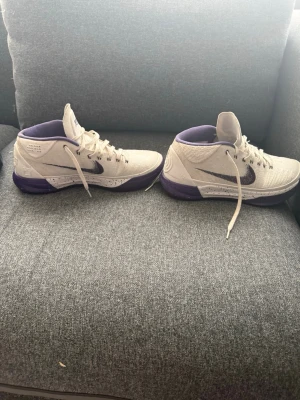 Nike Kobe vita och lila sneakers - Säljer ett par Nike Kobe sneakers i vitt och lila med klassisk Kobe-logga på plösen och signaturdetalj. Skorna har en mellanhög siluett, snörning och en robust sula i lila. Perfekta för dig som gillar basketstil och vill sticka ut med snygga detaljer. Inga sulor och ingen box
