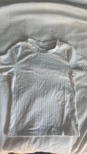 Vit ribbad t-shirt med struktur - Säljer en vit ribbad t-shirt med korta ärmar och rund hals. T-shirten har ett diskret randigt mönster i strukturen som ger en snygg och modern look. Perfekt till jeans eller kjol för en clean stil. Andvänd för länge sedan några gånger. Från Lindex storlek 134/140. Passar nog nån storlek större. Skriv om ni har frågor!