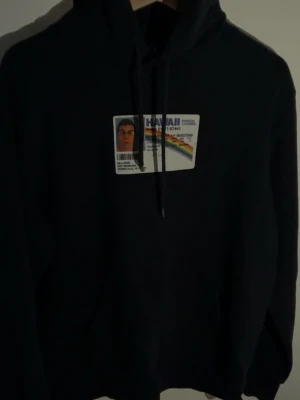 Marinblå hoodie McLovin Hawaii-tryck - Marinblå hoodie med huva och dragsnören. På bröstet finns ett stort tryck av ett Hawaii-leg med regnbåge och texten 'McLovin'. På ryggen står det 'McLovin' i vit skrivstil. Perfekt för dig som gillar filmreferenser och streetwear. Pris kan diskuteras.