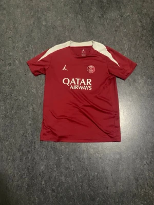 Röd PSG träningströja Dri-Fit - Säljer en röd PSG träningströja med vita detaljer och Qatar Airways-tryck på bröstet. Tröjan har korta ärmar och är tillverkad i ett lätt och snabbtorkande Dri-Fit material, perfekt för träning eller fotbollsmatcher.