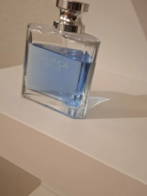 Nautica blå parfymflaska - Snygg parfym från Nautica i en stilren, fyrkantig glasflaska med silverfärgad kork. Parfymen har en fräsch blå färg som ger en modern känsla. Perfekt att ha framme på hyllan som en snygg detalj i badrummet.
