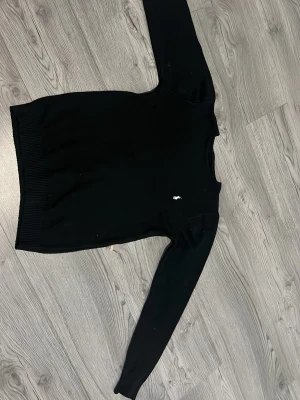 Svart stickad tröja från Ralph Lauren - Klassisk svart stickad tröja från Ralph Lauren med rund halsringning och den ikoniska vita broderade loggan på bröstet. Tröjan har långa ärmar och ribbade muddar vid ärmslut och nederkant. Perfekt till en stilren och enkel look.
