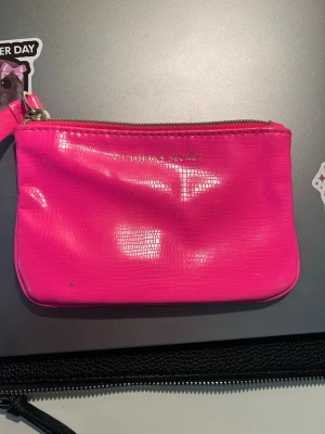 Rosa plånbok Victoria's Secret - Snygg och trendig plånbok i starkt rosa från Victoria's Secret. Plånboken har en glansig yta med subtilt mönster, dragkedja i guld och en liten handledsrem. Perfekt för kort, cash och smågrejer när du är på språng.