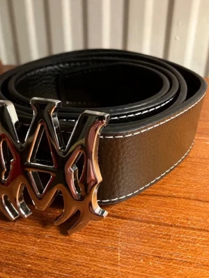 Svart bälte AMIRI - Snyggt svart bälte från AMIRI i skinn med vit söm längs kanterna och ett stort, unikt metallspänne i silverfärg. Perfekt för att ge outfiten en edgy touch och dra blickarna till sig. Bältet har en bred design och känns riktigt trendigt. Helt ny dm om ni undrar nått, kommer med hålgörare