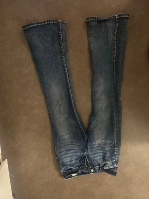 Mörkblå bootcut jeans med broderade fickor - Säljer ett par mörkblå bootcut jeans med snygga slitningar och broderade bakfickor med vita detaljer. Jeansen har klassisk femficksmodell och är tillverkade i denim med lätt stretch. Perfekta för dig som gillar en avslappnad men trendig look.