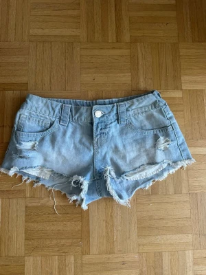 Ljusa jeansshorts med slitningar - Säljer ett par ljusblå jeansshorts med råa kanter och slitna detaljer framtill. Shortsen har fem fickor, bälteshällor och knappstängning. Perfekta för varma dagar och ger en avslappnad look.