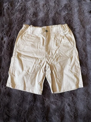 Beige bomullsshorts med fickor - Snygga beige shorts i bomull med klassisk passform. Shortsen har två sidofickor fram och en bakficka, samt bälteshällor och knappstängning. Perfekta för varma dagar och enkel att matcha med olika stilar.