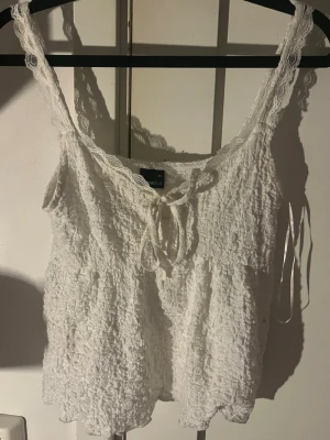 Vit spetslinnetopp från Gina Tricot - Söt vit linnetopp från Gina Tricot i storlek XS. Toppen har spetsdetaljer vid axelbanden, knytning fram och en krinklad struktur. Perfekt för sommarens alla outfits och ger en romantisk vibe.
