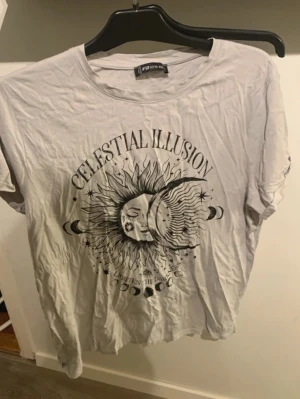 Vit t-shirt med tryck Celestial Illusion - Vit t-shirt med svart tryck av sol, måne och texten 'Celestial Illusion'. Rund halsringning och korta ärmar. Mjuk och skön, perfekt för en avslappnad stil.