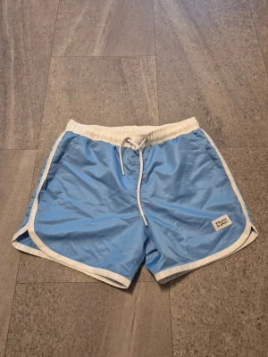 Ljusblå nya badshorts från Frank Dandy - Snygga ljusblå badshorts från Frank Dandy med vita detaljer och snörning i midjan. Shortsen är använda endast en gång och har bra passform , i storlek medium.