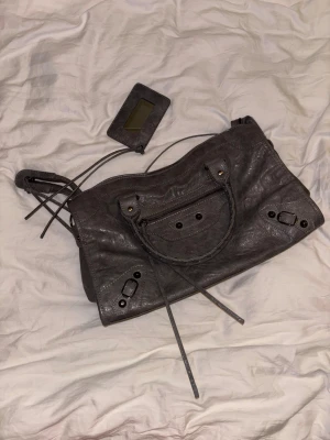 Grå handväska med detaljer - Snygg grå handväska i mjukt läder med flätade handtag och dekorativa nitar. Väskan har dragkedja framtill, långa läderband och en matchande spegelaccessoar. Något ljusare i verkligheten och aldrig använd!