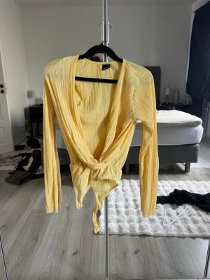 Gul plisserad body från Gina Tricot - Trendig gul body från Gina Tricot i plisserat material. Långärmad modell med djup omlott-ringning och knyt framtill. Perfekt för dig som vill ha en unik och snygg detalj i garderoben. Materialet är lätt och stretchigt för skön passform.