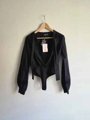Ny Missguided body i blusmodell - tags kvar - Superfin och elegant svart body från Missguided (köpt på Nelly). Helt oanvänd – lapp/tagg sitter fortfarande kvar! 🏷️  Feminin omlottdesign med djup V-ringning, dekorativa band som knyts runt halsen och fina spets-/meshdetaljer vid ärmsluten. Den blusliknande modellen ger en dressad och elegant look ✨  Perfekt till fest, middag eller utekväll 🥂 men också supersnygg till jeans eller kjol för en mer uppklädd. 💌Hör gärna av dig vid frågor, fler bilder eller om mått önskas 