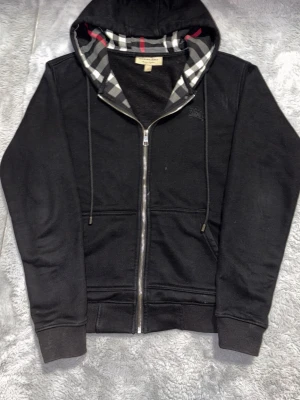 Svart Burberry hoodie med dragkedja - Snygg svart hoodie från Burberry med dragkedja och klassiskt rutigt foder i huvan. Broderad logga på bröstet och två fickor framtill. Tillverkad i mjukt material som känns skönt mot huden. Perfekt för en avslappnad och stilren look.