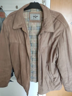 Beige mockajacka från Reportage - Säljer en beige mockajacka från Reportage med klassiskt rutigt innerfoder. Jackan har dragkedja framtill, två sidofickor och en innerficka. Stilig krage och mjukt material som ger en snygg och avslappnad look.