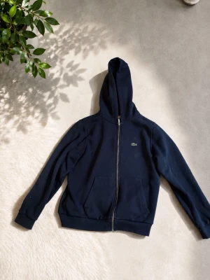Mörkblå hoodie från Lacoste - Snygg mörkblå hoodie från Lacoste med dragkedja framtill och klassisk krokodil-logga på bröstet. Hoodien har två stora fickor fram och en skön huva. Tillverkad i mjukt bomullsmaterial som känns bekvämt hela dagen.