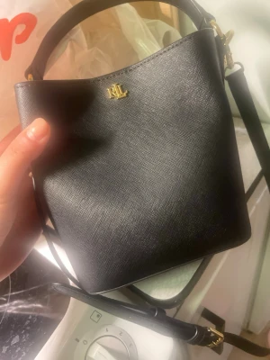 Svart bucket bag från Ralph Lauren - Stilren svart handväska i bucket-modell från Ralph Lauren med guldfärgade detaljer och RL-logga framtill. Väskan har både ett kort handtag och en längre axelrem, tillverkad i strukturerat läder för en lyxig känsla. Använts några gånger men ser fortfarande splitter ny ut. Inga skador på väskan samt man får med sig en dustbag också