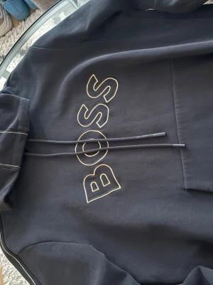 BOSS hoodie  - Svart hoodie från BOSS med stor guldkantad logga på bröstet. Klassisk huva med dragsko och synliga sömmar på ärmarna. Mjuk bomullskänsla och känguruficka framtill. Perfekt för en stilren och avslappnad look.
