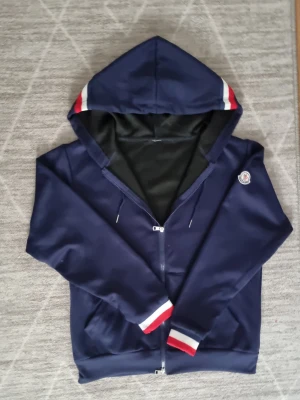 Marinblå Moncler hoodie med dragkedja - Snygg marinblå hoodie från Moncler med dragkedja, huva och vita/röda detaljer på muddar och huva. Klassisk Moncler-logga på ärmen. Tillverkad i mjukt material som känns skönt mot huden. Perfekt för en avslappnad och trendig look.