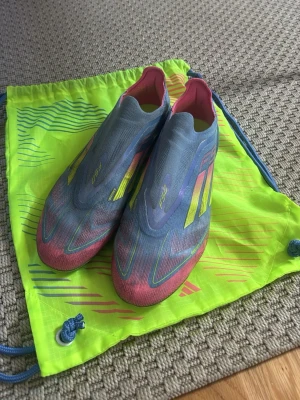 Adidas F50 blå/rosa fotbollsskor - Säljer ett par Adidas F50 AG fotbollsskor. Köpta på Unisport för 3199 kr. Tillverkade i lätt syntetmaterial för maximal fart på planen. Medföljer original gympåse i neonfärg, samt skohorn.